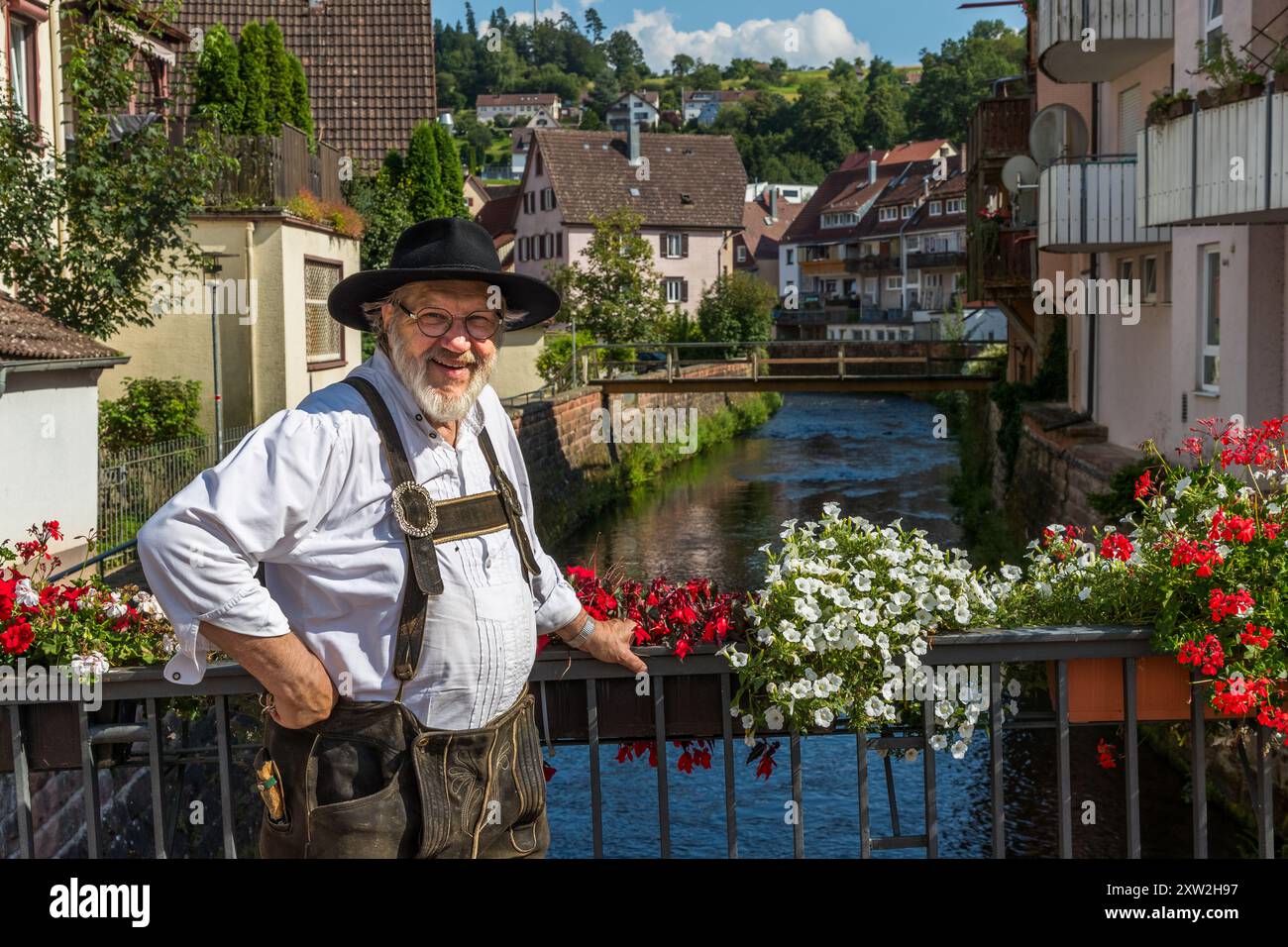 Raftsman Martin Spreng stands in traditional dress on a bridge over the Nagold river on Rosenstraße in Altensteig, Baden-Württemberg, Germany. Red and white flowers decorate the bridge railing, with the historic old town lining the river behind him. Spreng chairs the German Rafting Association. Rafting has been recognised as UNESCO intangible cultural heritage since 2022. Stock Photo