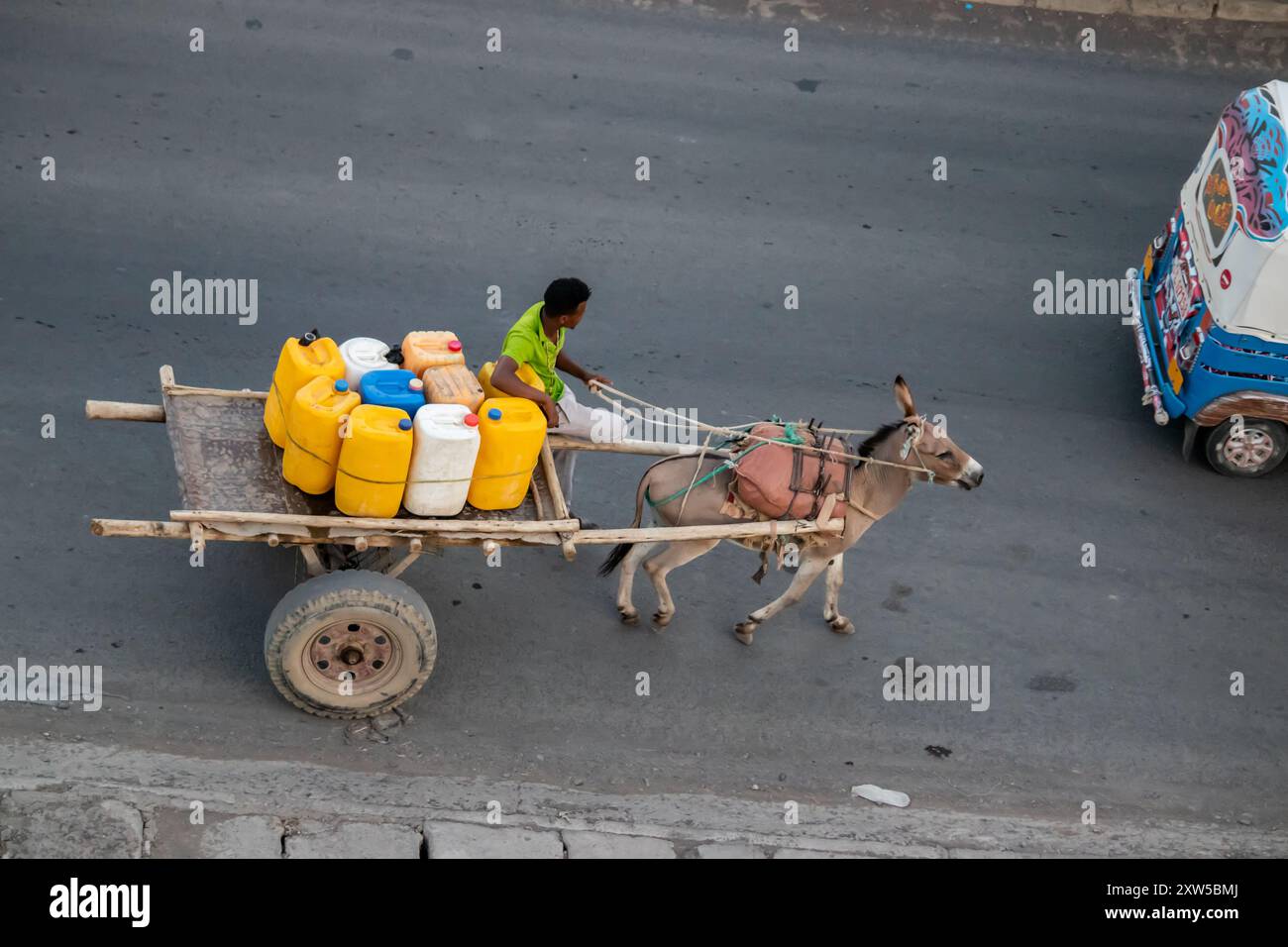 In rural Africa, a young man runs a business distributing water in colorful jerrycans, using a primitive wooden trailer pulled by a donkey Stock Photo