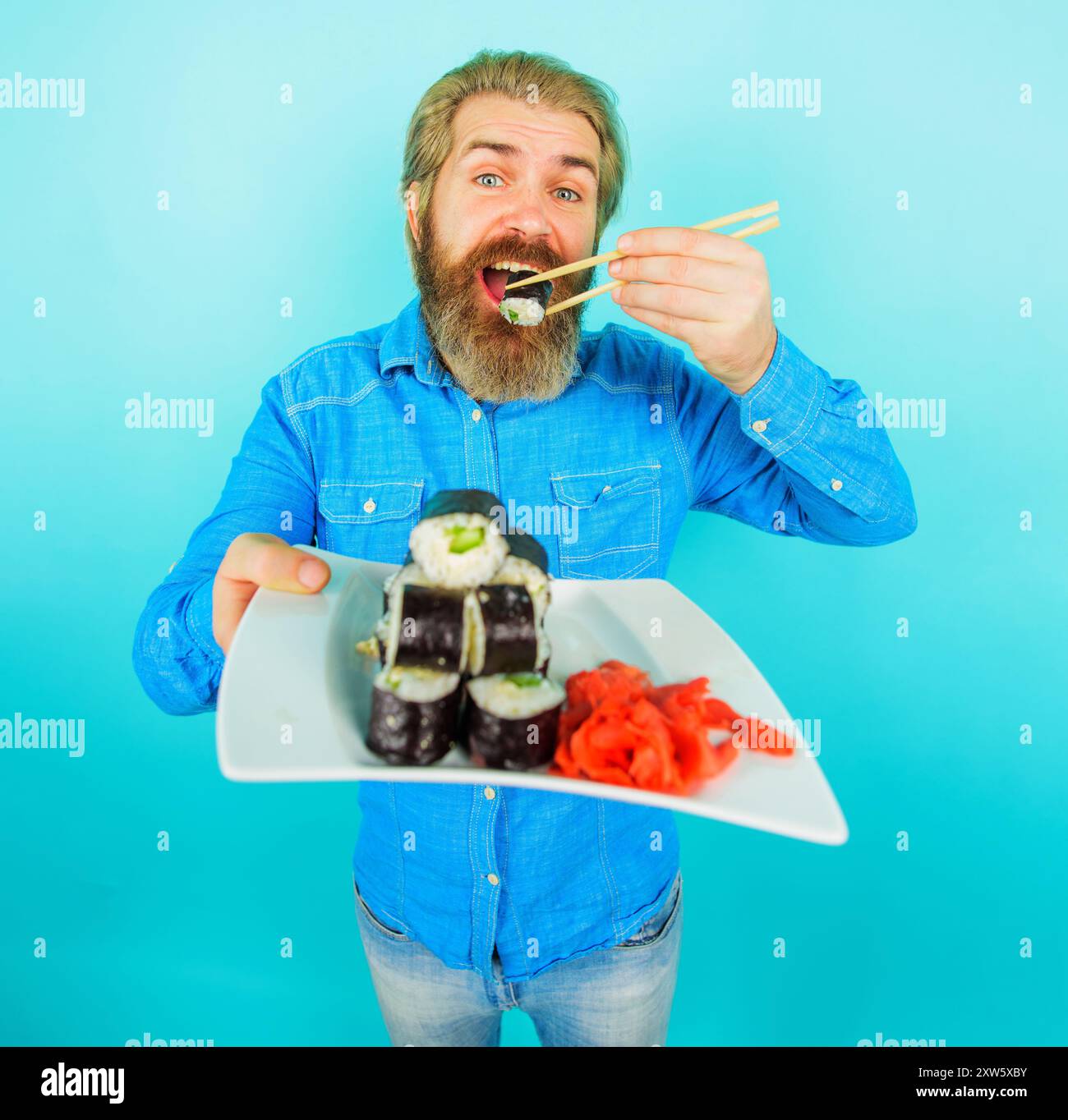 Sushi delivery or takeaway food concept. Bearded man eating tasty sushi roll with chopsticks enjoying traditional japanese food. Smiling man with plat Stock Photo