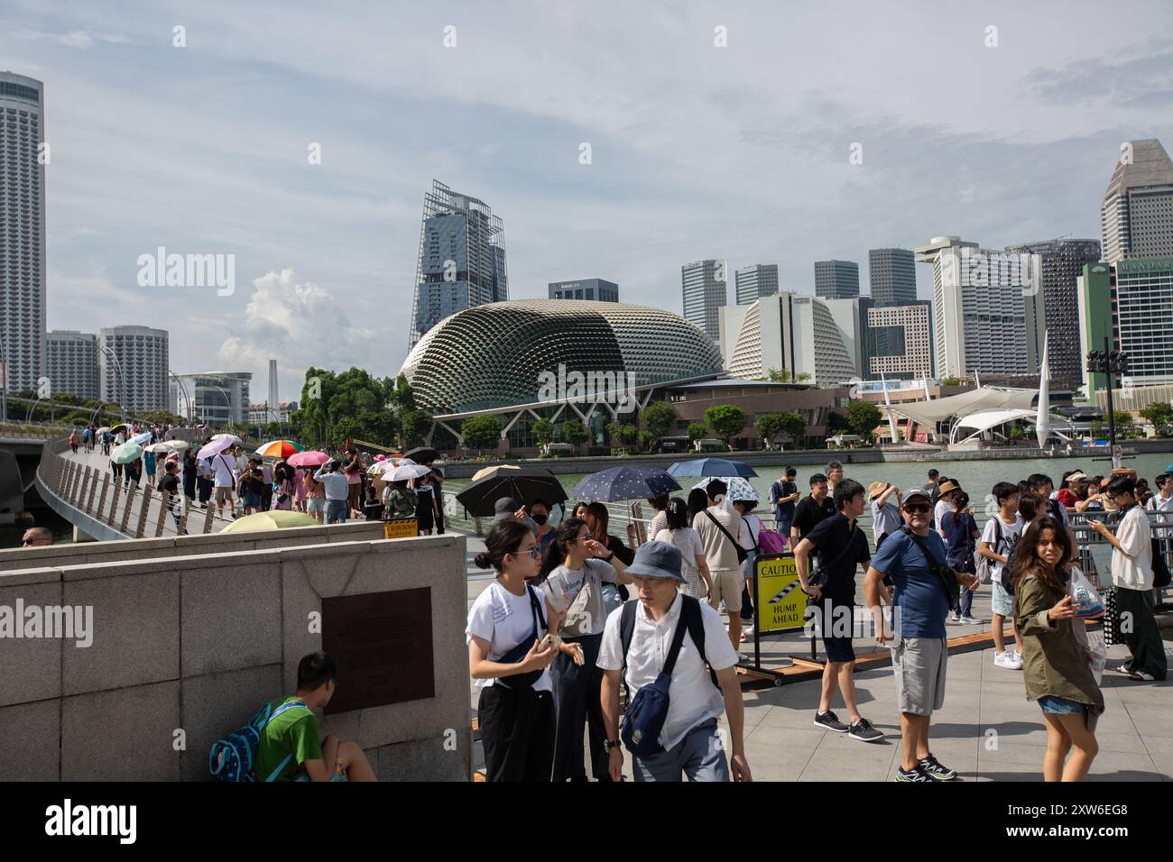 With climate change impact. People tourists seen holding umbrella, wear hat to brave the hot sun heat at Merlion Park while sightseeing. Singapore. Stock Photo