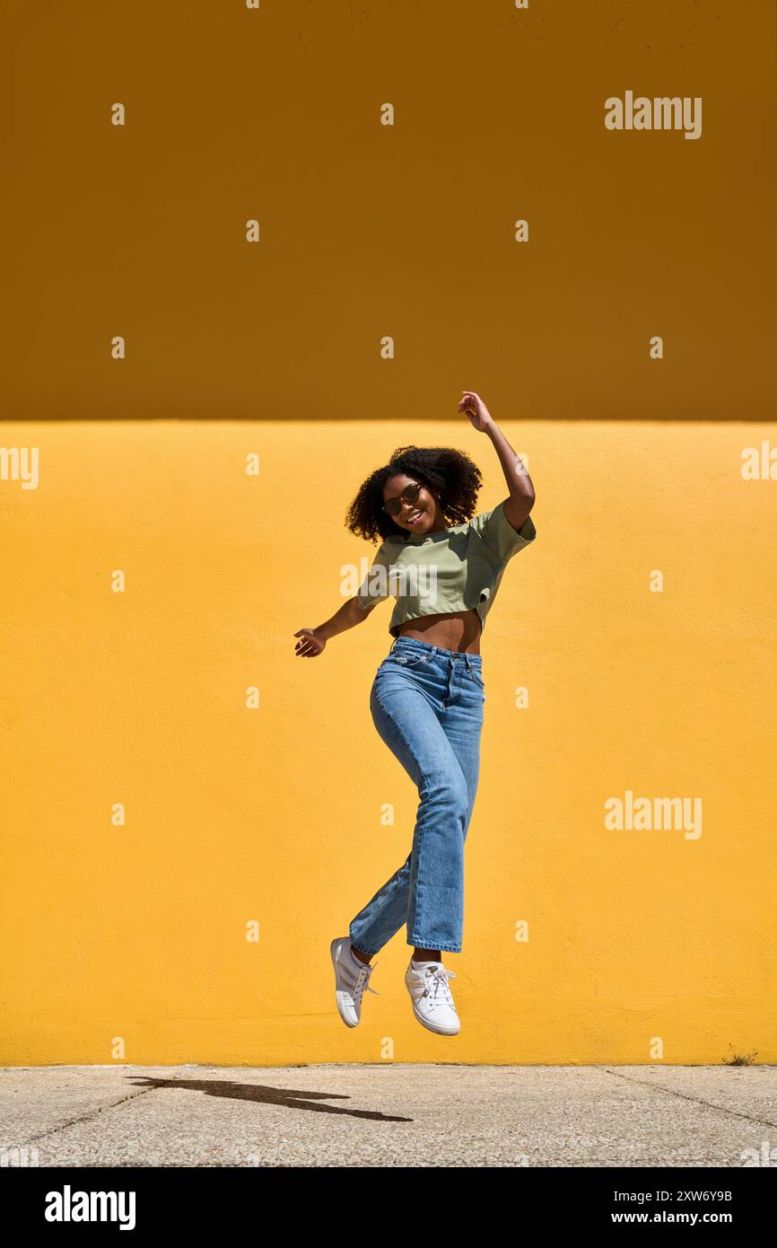 Happy funky young African American woman winner jumping at yellow wall ...