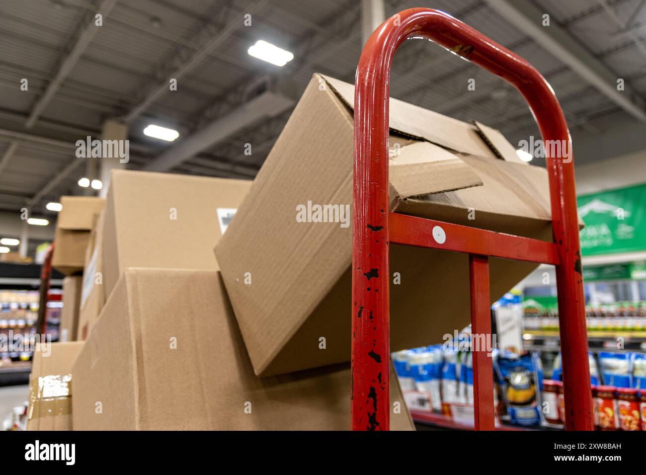 Red hand truck loaded with cardboard boxes in a warehouse aisle - flanked by shelves stocked with various products. Taken in Toronto, Canada. - Stock Image