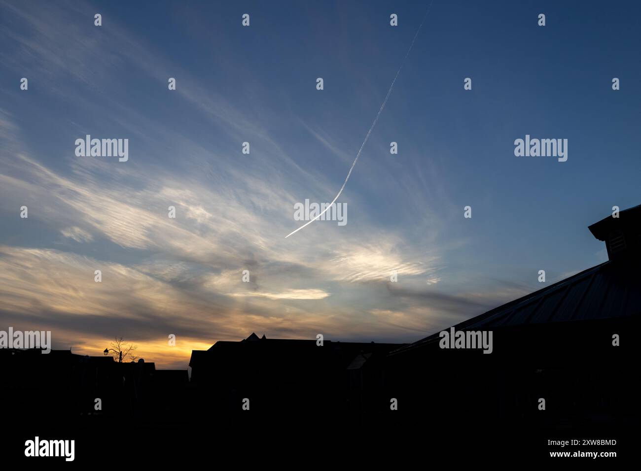 Sunset silhouette of a suburban skyline with contrail - vibrant blue and orange sky streaked with wispy clouds. Taken in Toronto, Canada. - Stock Image