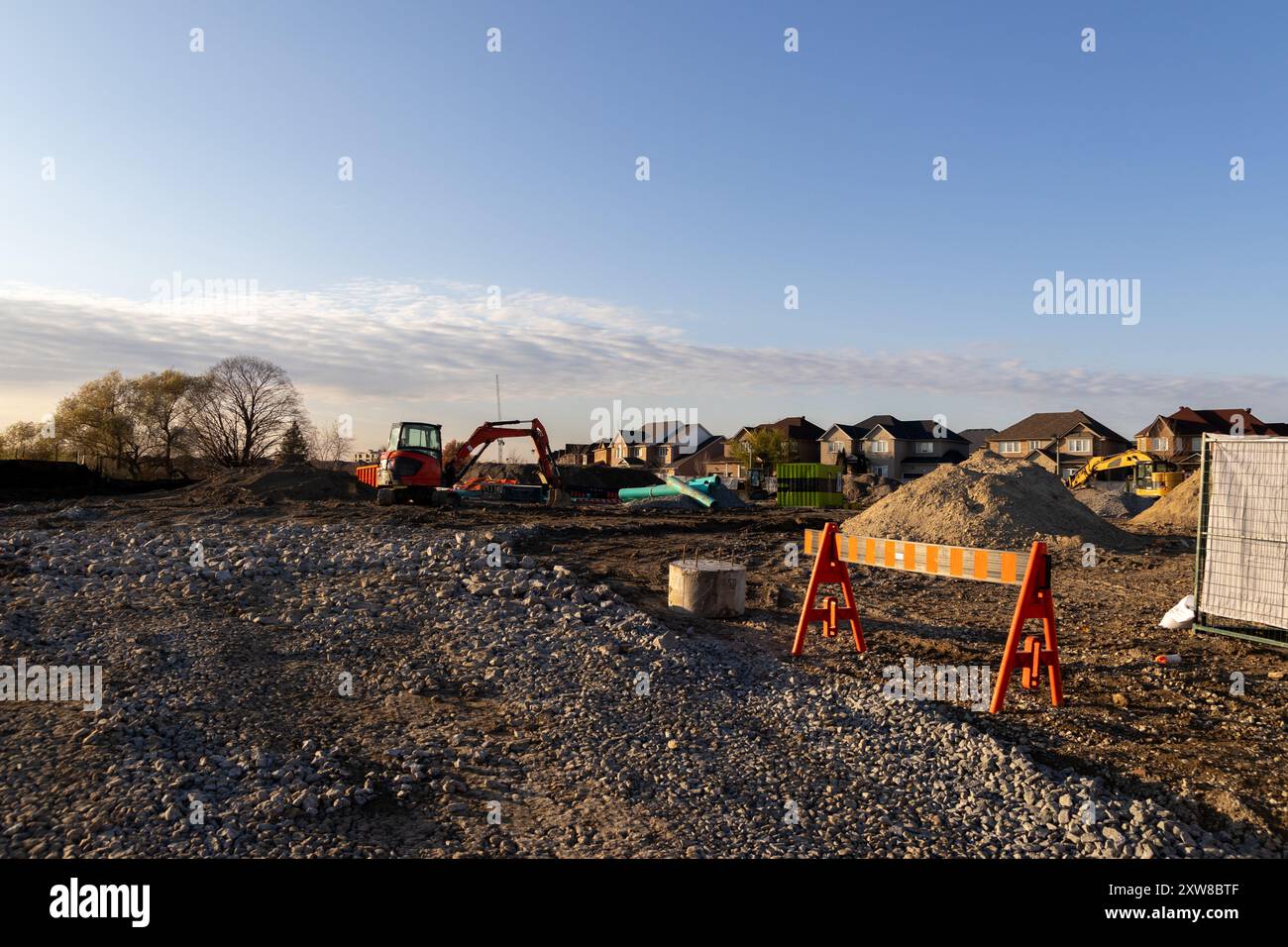 Sunset-lit construction site with red excavator and scattered building materials - suburban neighborhood backdrop. Taken in Toronto, Canada. - Stock Image