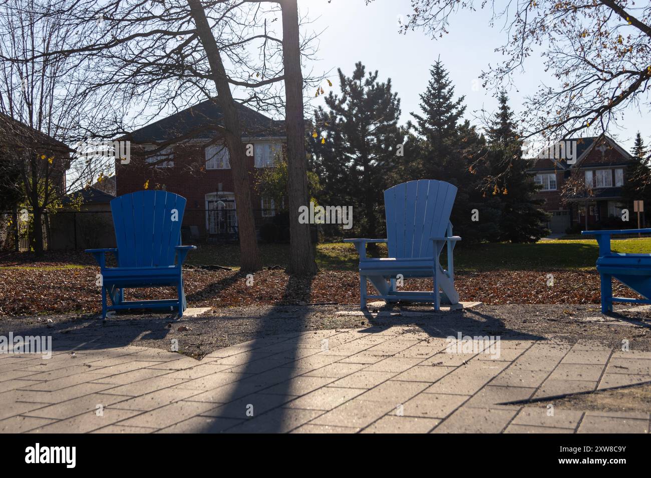 Bright autumn day with vibrant blue Adirondack chairs on a patio, casting long shadows with suburban homes and bare trees in background. Taken in Toro - Stock Image