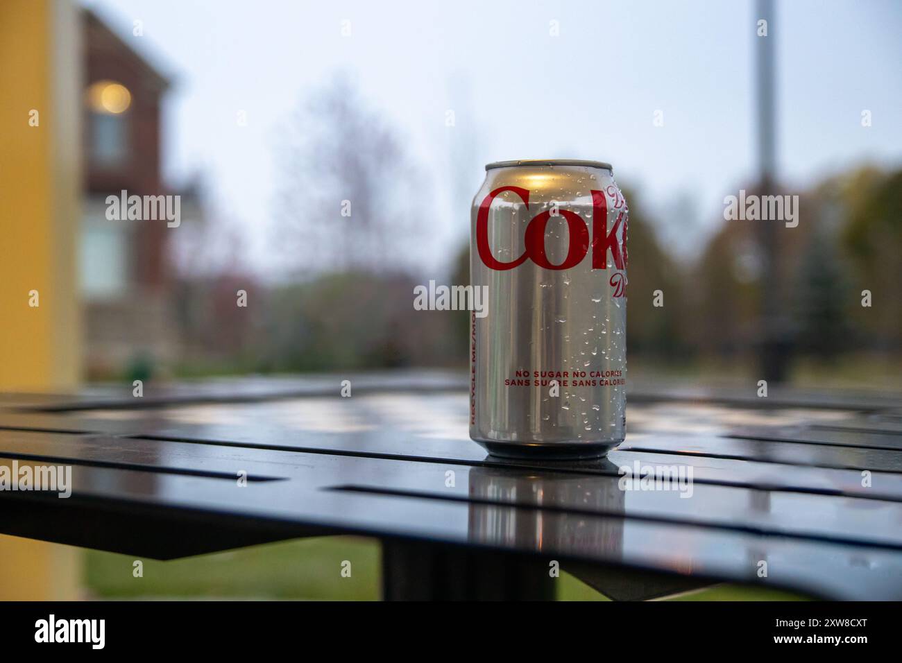 Aluminum can of Coca-Cola Zero Sugar with condensation - resting on outdoor metal table - blurred background featuring trees and a building at dusk. T - Stock Image