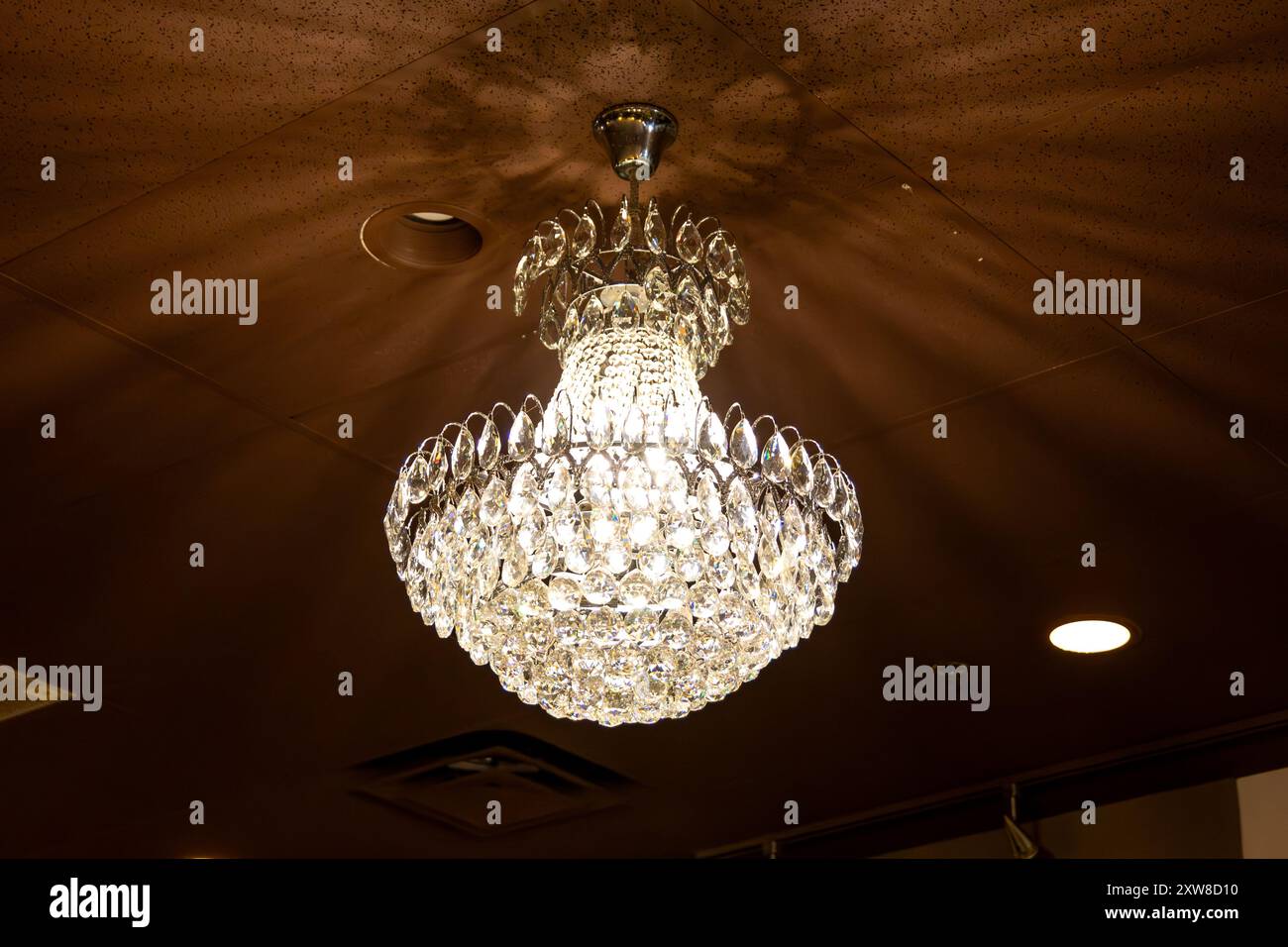 Elegant crystal chandelier hangs from a textured ceiling, casting a warm glow - intricate glass detailing. Taken in Toronto, Canada. - Stock Image