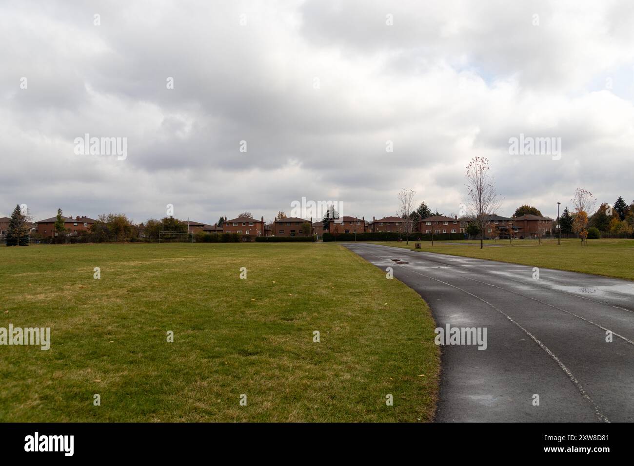 Overcast sky above a winding park path leading towards suburban houses, flanked by expansive green lawns and sparse autumn trees. Taken in Toronto, Ca - Stock Image