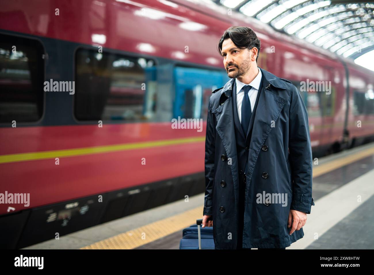 Confident businessman in stylish suit waits at train station, clutching suitcase tightly, exuding seriousness and thoughtfulness Stock Photo