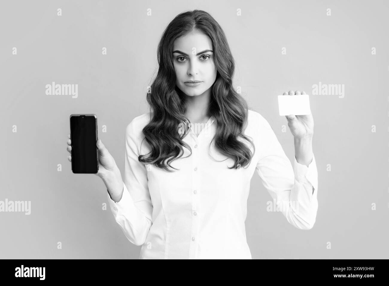 Amazed young brunette woman in shirt using mobile cell smart phone hold credit bank card isolated on gray background studio portrait. Stock Photo