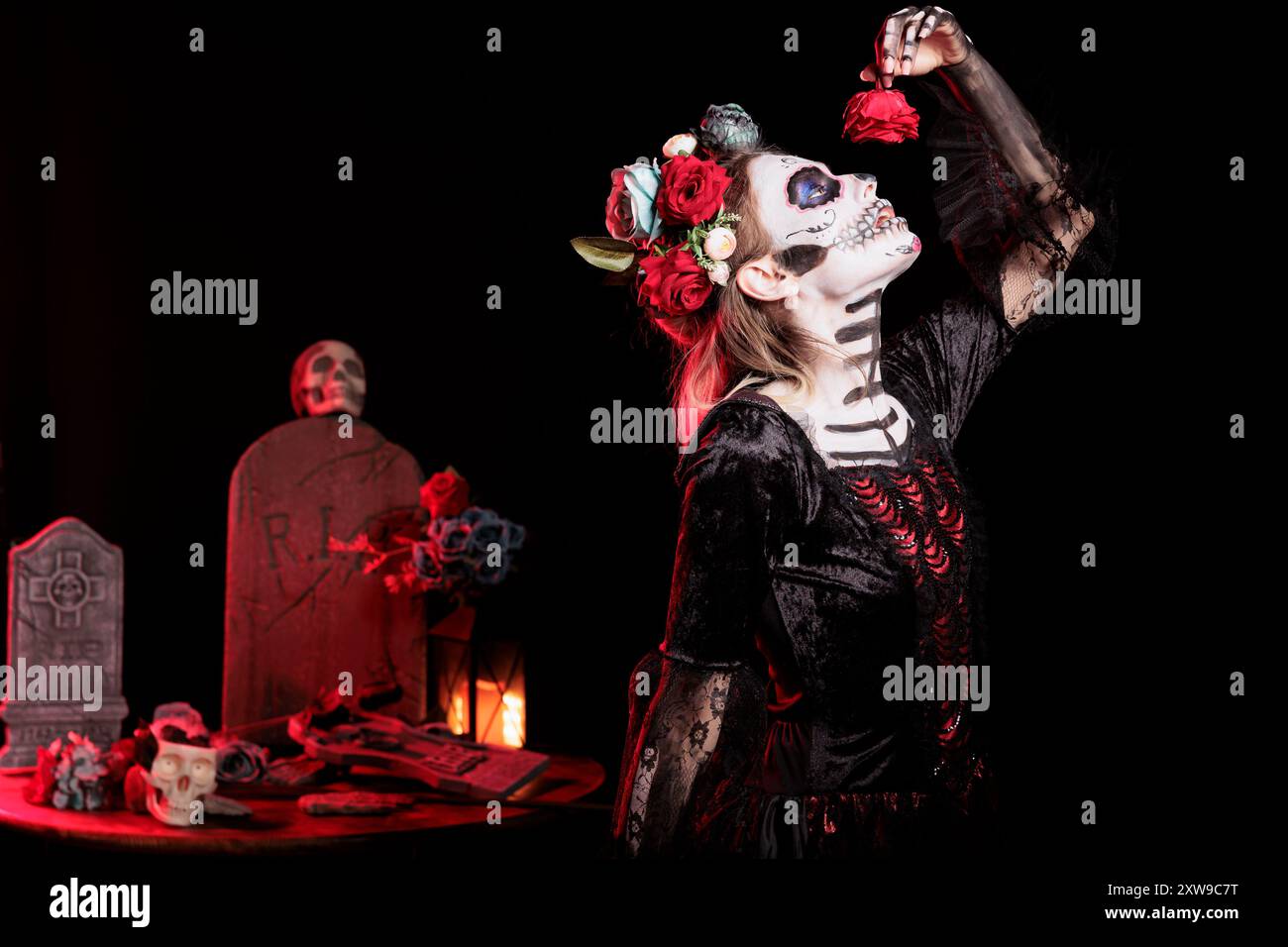 Woman posing for Day of the Dead Mexican celebration honoring dead loved ones, pretending to eat red rose. Goddess lady doing ancient Aztec custom of celebrating ancestors, devouring flower Stock Photo