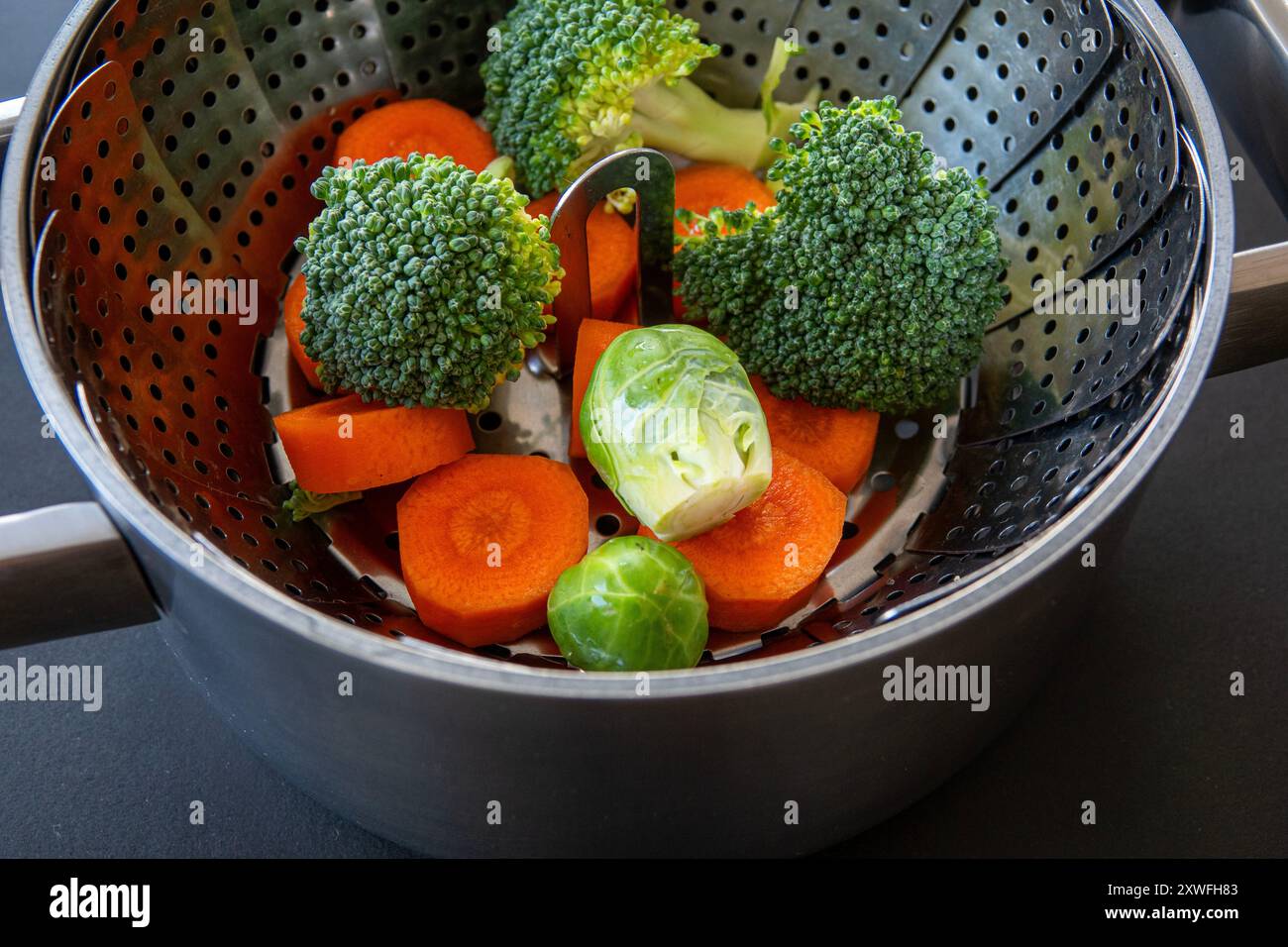 Collapsible food steaming basket in a pot with broccoli, carrots and Brussels sprouts. Steaming helps retain the essential nutrients and flavour and h Stock Photo