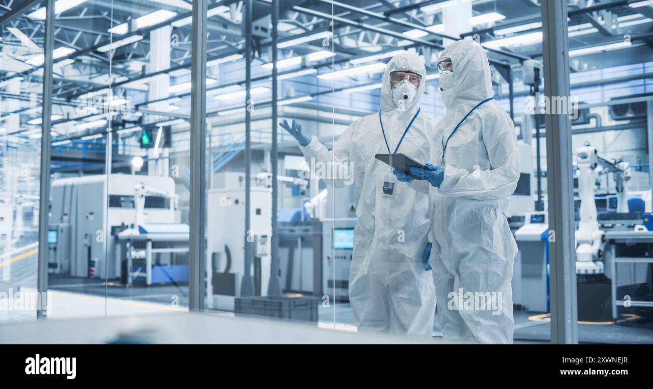 Development Factory Cleanroom: Engineers Wearing Coveralls and Goggles while Talking and Using Tablet Computer. Scientists Working in Advanced Electronics Manufacture with Precision Machinery Stock Photo
