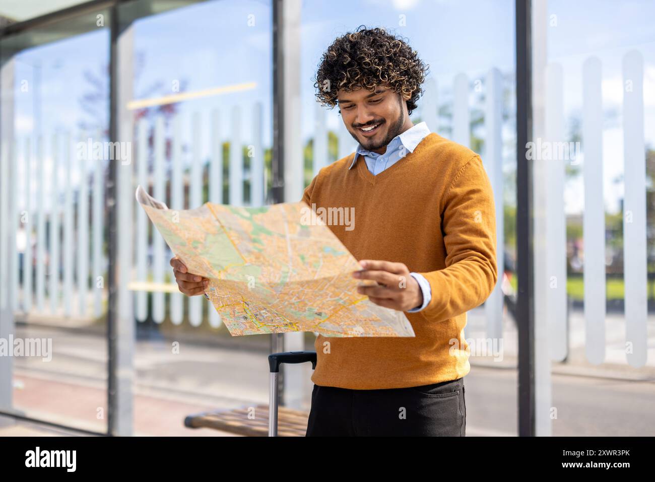 Happy young man with curly hair exploring city using map, standing at bus stop with suitcase. Captures travel, adventure, and navigation on bright sunny day. Stock Photo