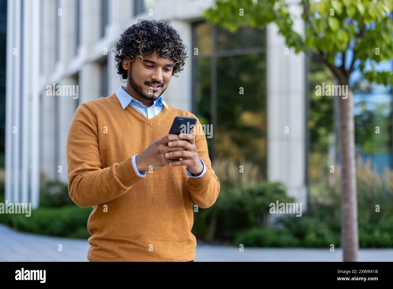 Young man enjoying outdoor work break, standing with phone in hand. Wearing casual sweater, he engages with technology in urban environment surrounded by nature, reflecting modern lifestyle Stock Photo