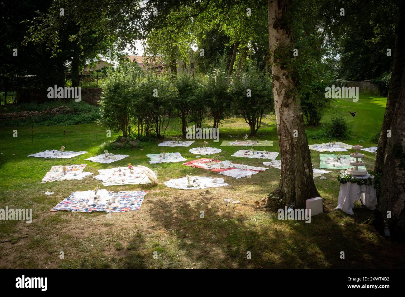 A beautiful outdoor wedding celebration featuring multiple picnic blankets spread out in a picturesque garden. Guests are seen enjoying the serene atmosphere, symbolizing unity and togetherness in nature. Stock Photo