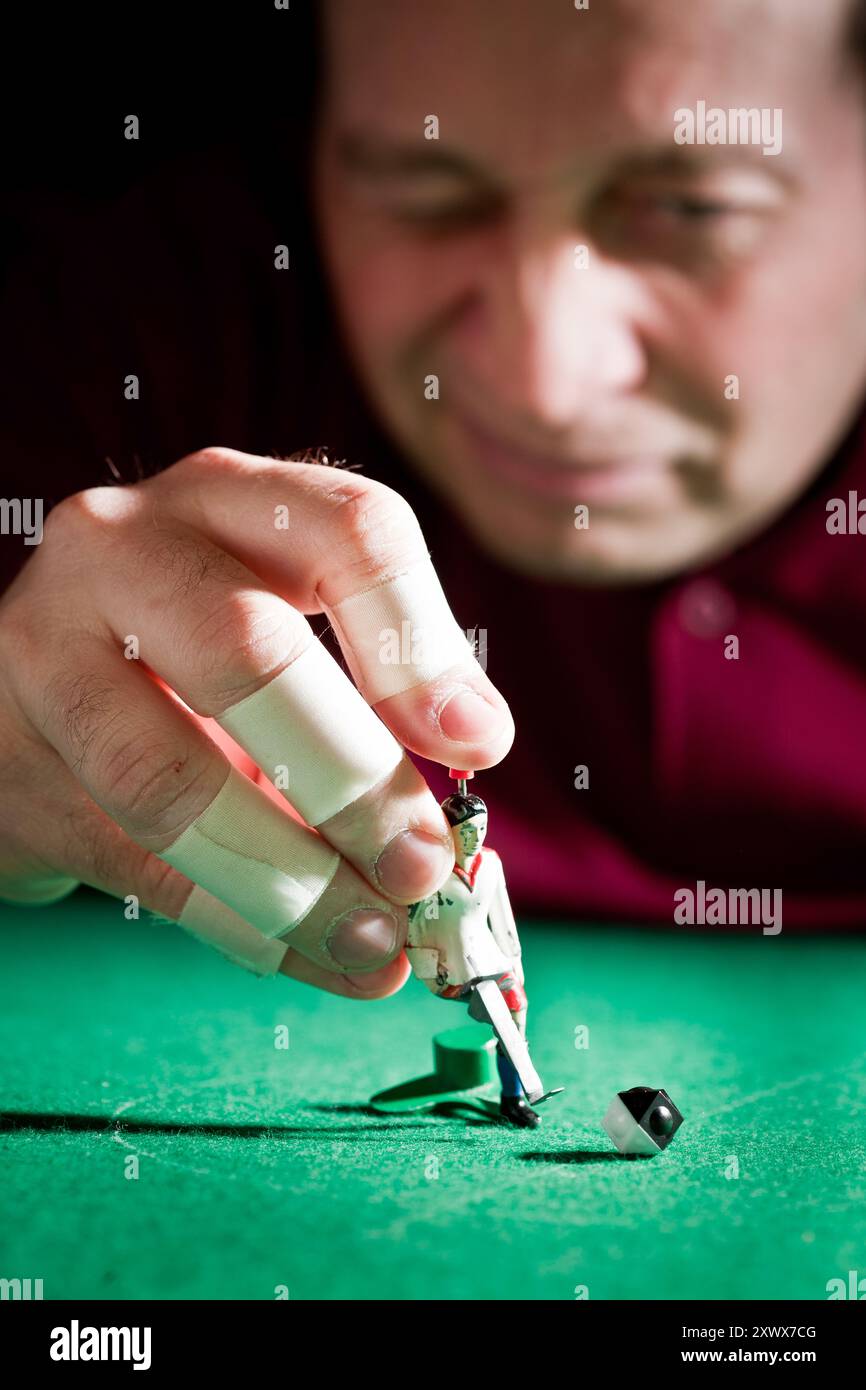 Close-up of a hand with tape on fingers playing Tipp-Kick, focusing on the miniature football player and ball on a green field. Captures the essence of table football games. Stock Photo