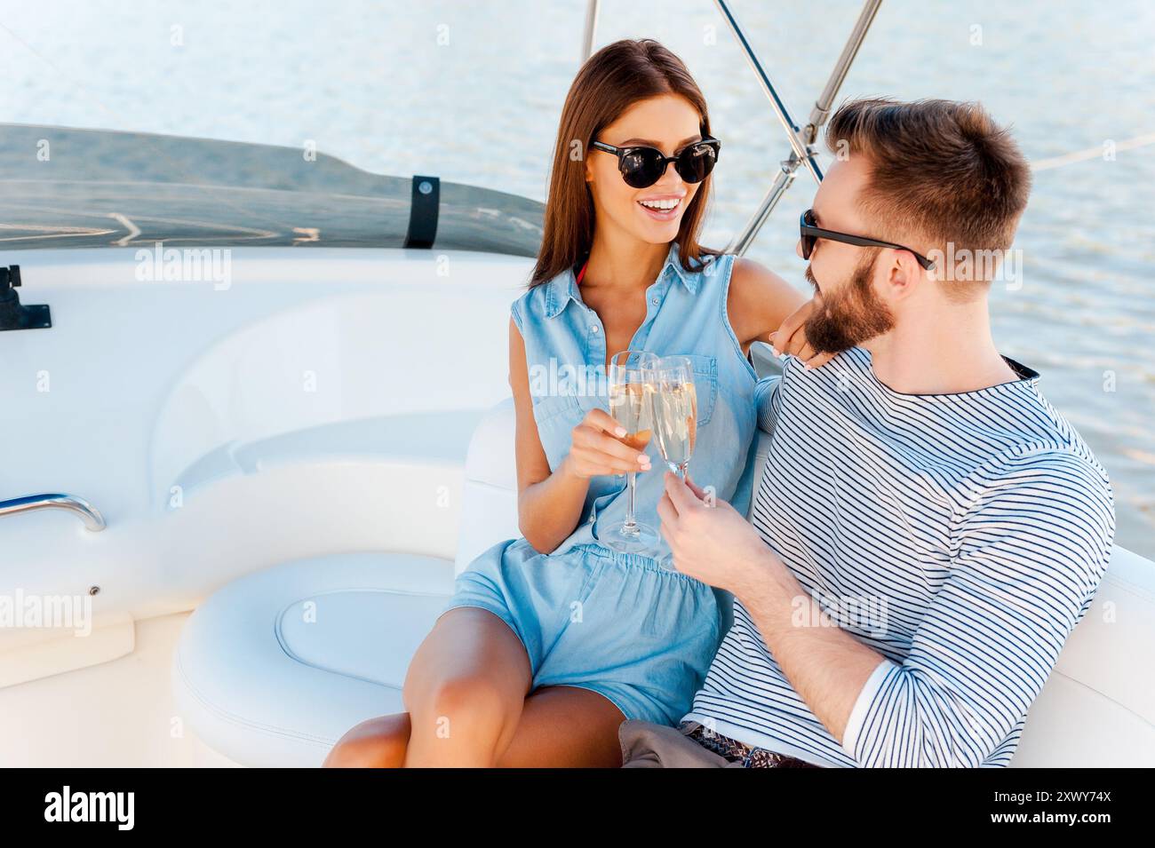 Celebrating their special anniversary. Smiling young couple holding glasses with champagne and looking at each other while sitting on the board of yac Stock Photo