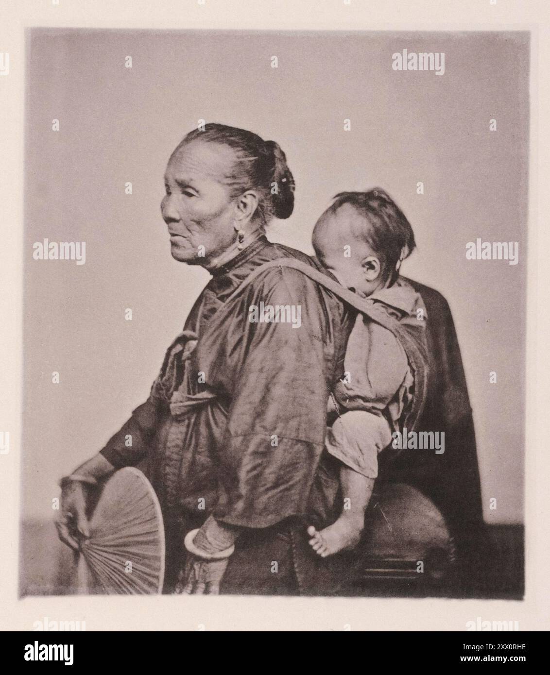 Vintage photo of an old Canton boat-woman and child. Illustrations of China, 1875 Many thousands of the population of Canton pass their lives in their boats; - in them they are born; and from them they are carried to their graves.; The old woman in the photograph is the grandmother living with her son's family in the boat; she still works cheerfully at the oar to help the domestic earnings; and nurses; all the while; one of the grandchildren. The babe is carried in a cloth slung over the shoulders; after the manner of the Chinese race. Stock Photo