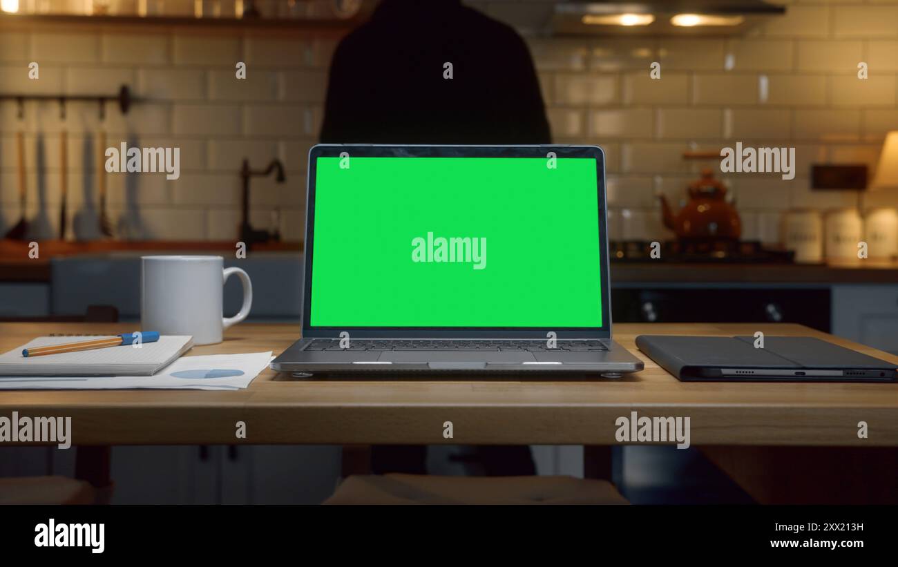 Laptop computer with mock up green screen chromakey display standing on the table. Man pouring hot water or tea to mug in the background in kitchen at Stock Photo