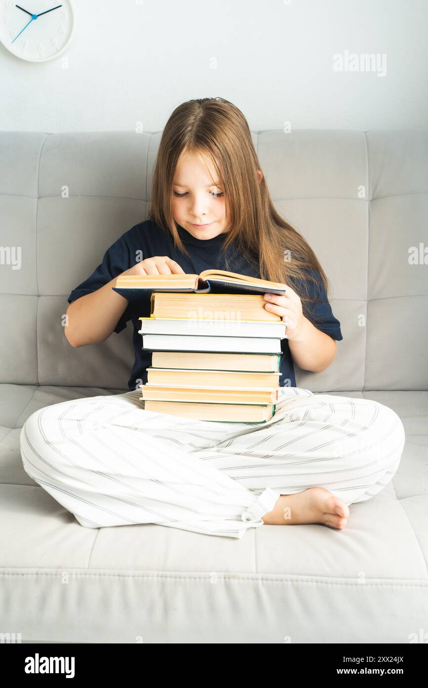 Little pre-teen girl with a stack of books reading on gray sofa at home ...