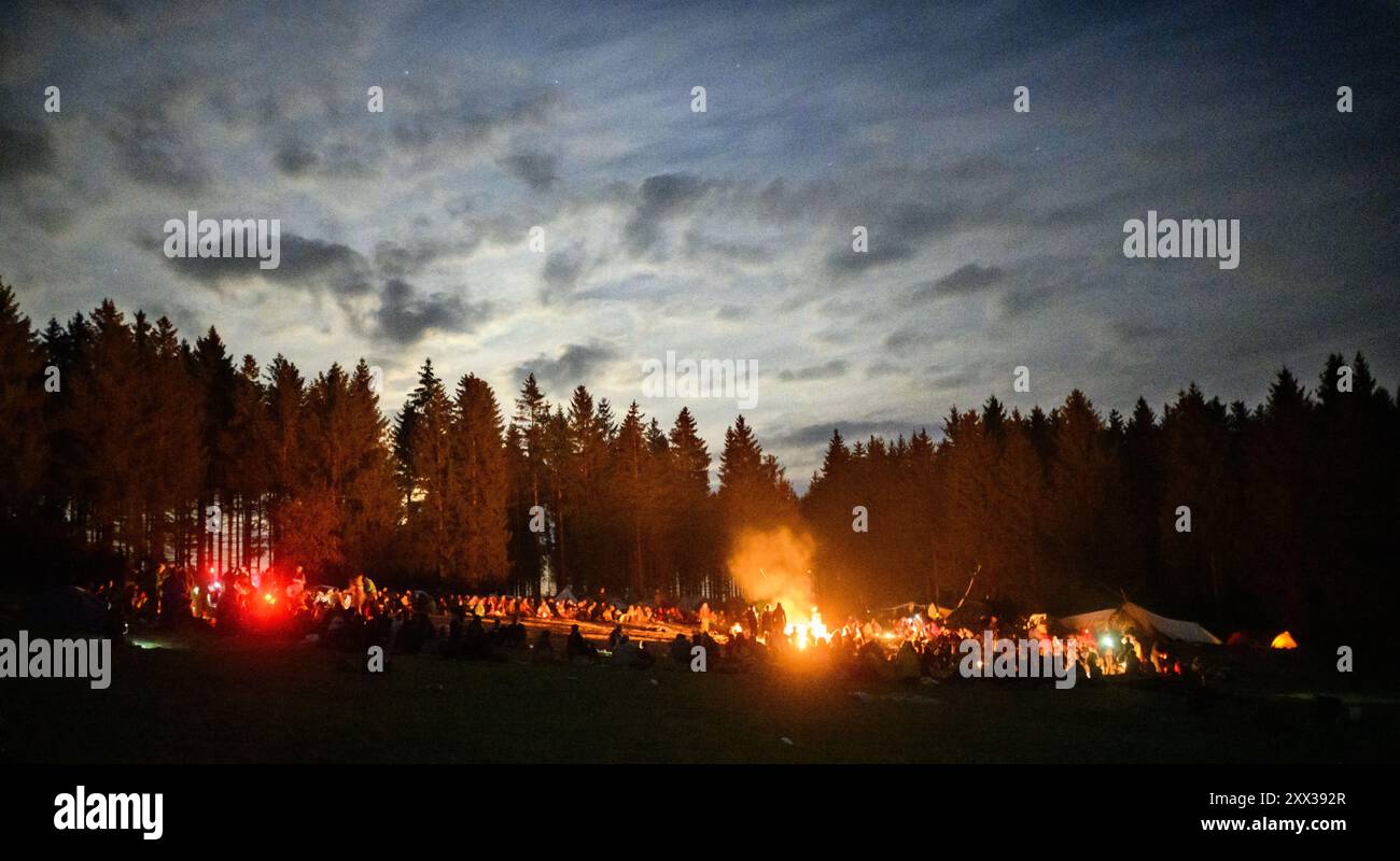 Bad Grund, Germany. 21st Aug, 2024. Hundreds of participants sit in a large circle around a campfire at a tent camp and eat the dinner they have prepared together. In the Harz Mountains, more than 1,000 people from all over the world are currently camping in the middle of the forest - albeit illegally. The campers of the 'Rainbow Family' movement, who are often simply referred to as 'hippies', are campaigning for world peace with their 'Rainbow Gathering' tent camp. Credit: Julian Stratenschulte/dpa/Alamy Live News Stock Photo