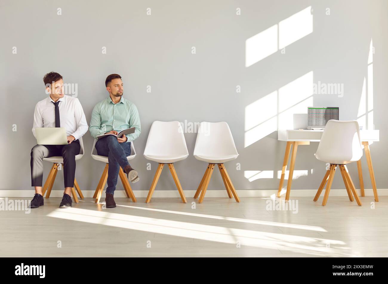 Two men sitting by office wall, waiting for job interview and looking to side at table with laptop Stock Photo