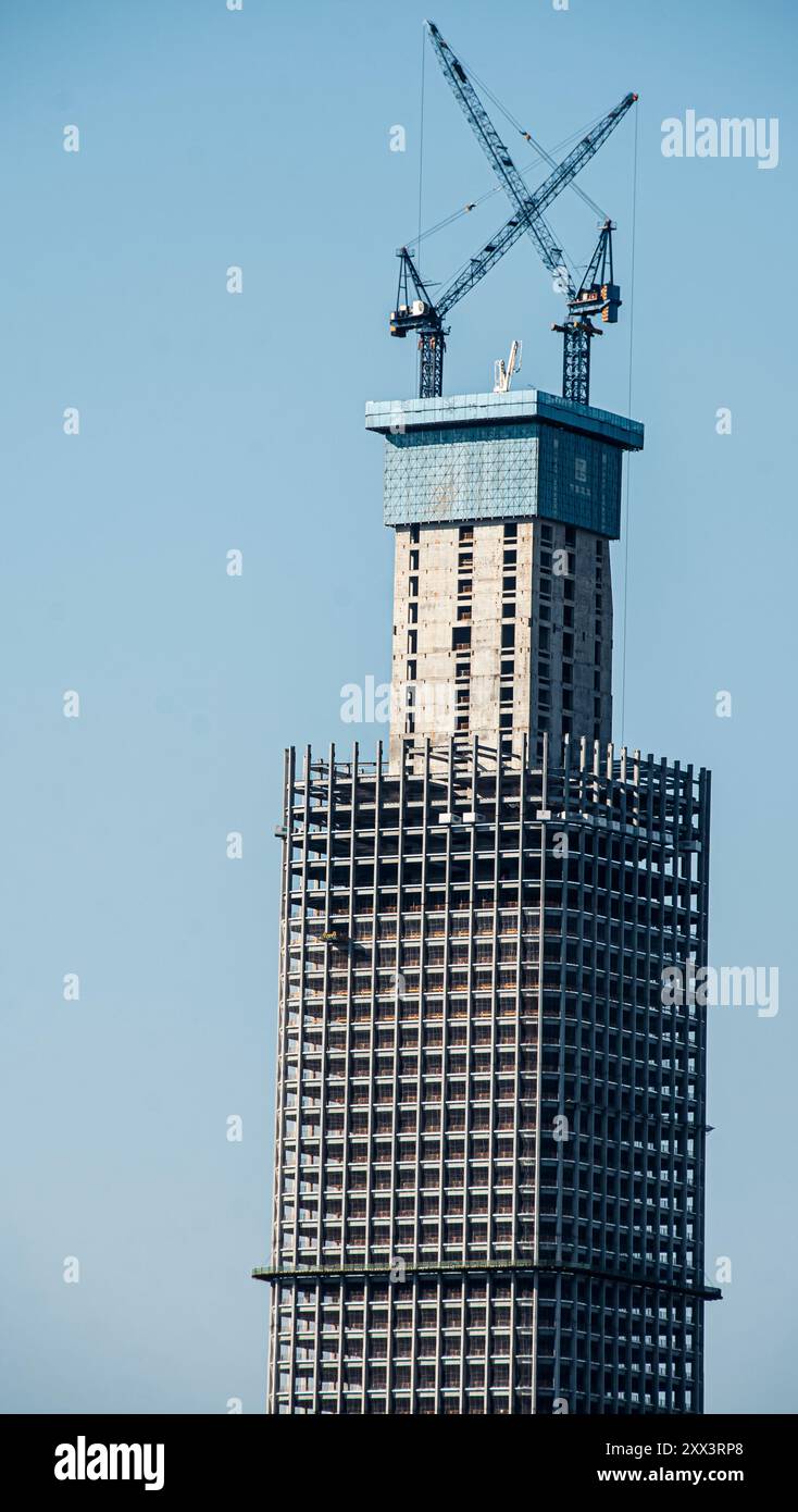 A tall skyscraper under construction with cranes on top against a clear ...