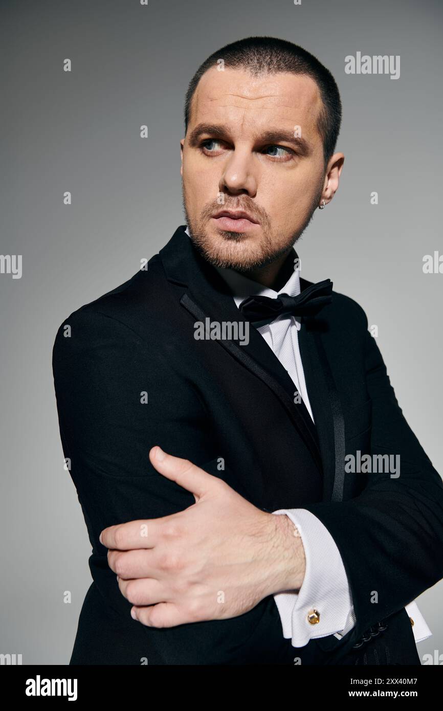 A stylish man in a black suit and bow tie stands against a grey ...