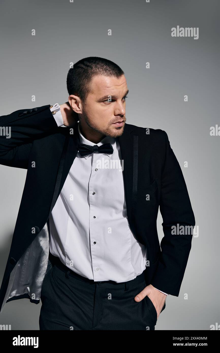 A man in a black suit and bow tie stands against a gray wall, adjusting ...