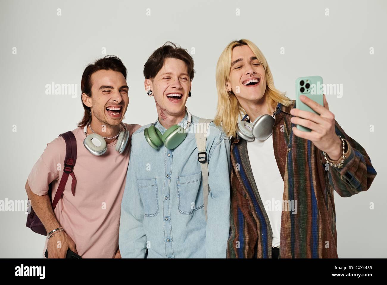 Three young, queer students laugh together while taking a selfie on a grey background. Stock Photo
