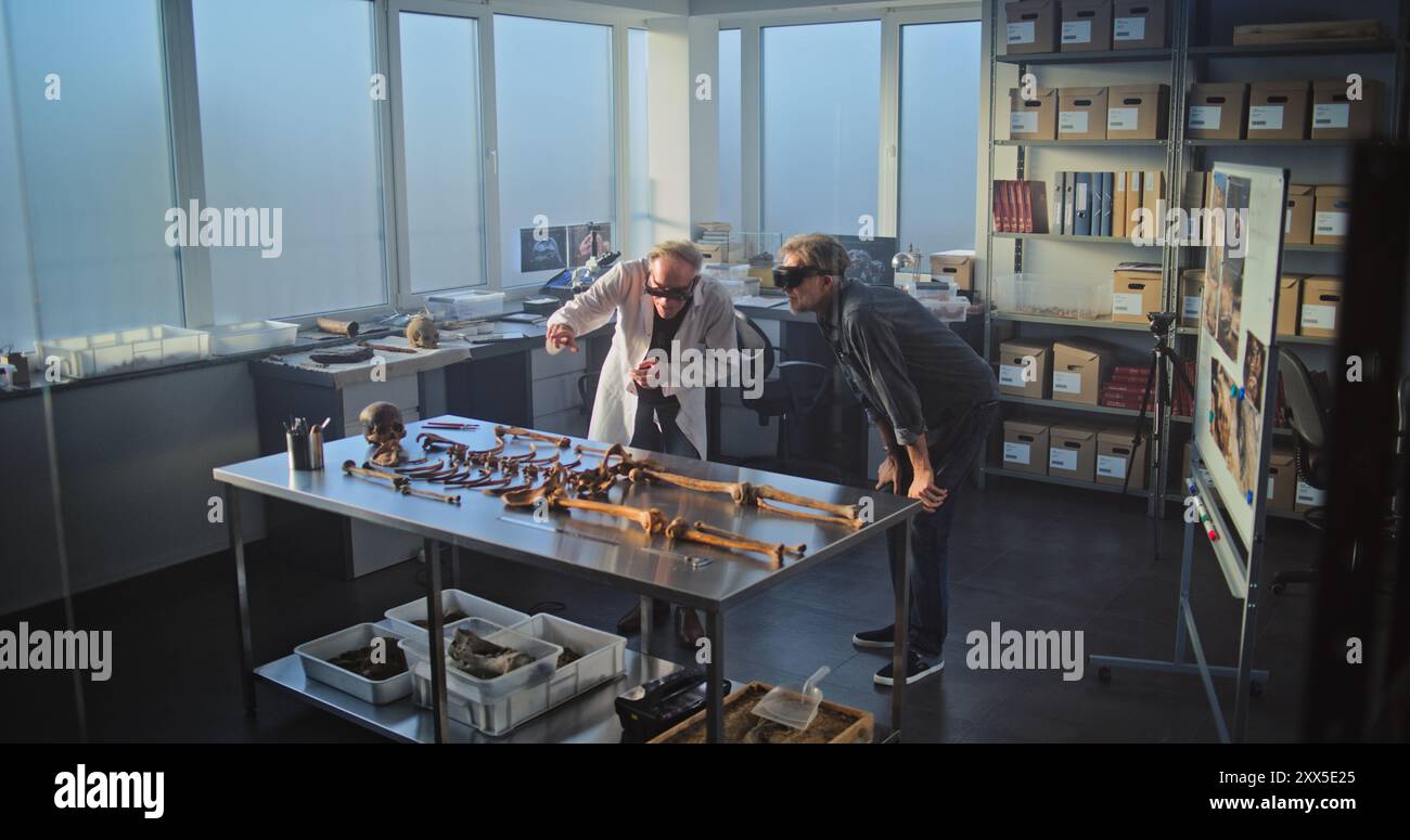 Two archaeologists conduct historical and scientific research in archaeological laboratory using virtual reality technology. Scientist in lab coat uses tablet computer, colleague wears AR headset. Stock Photo