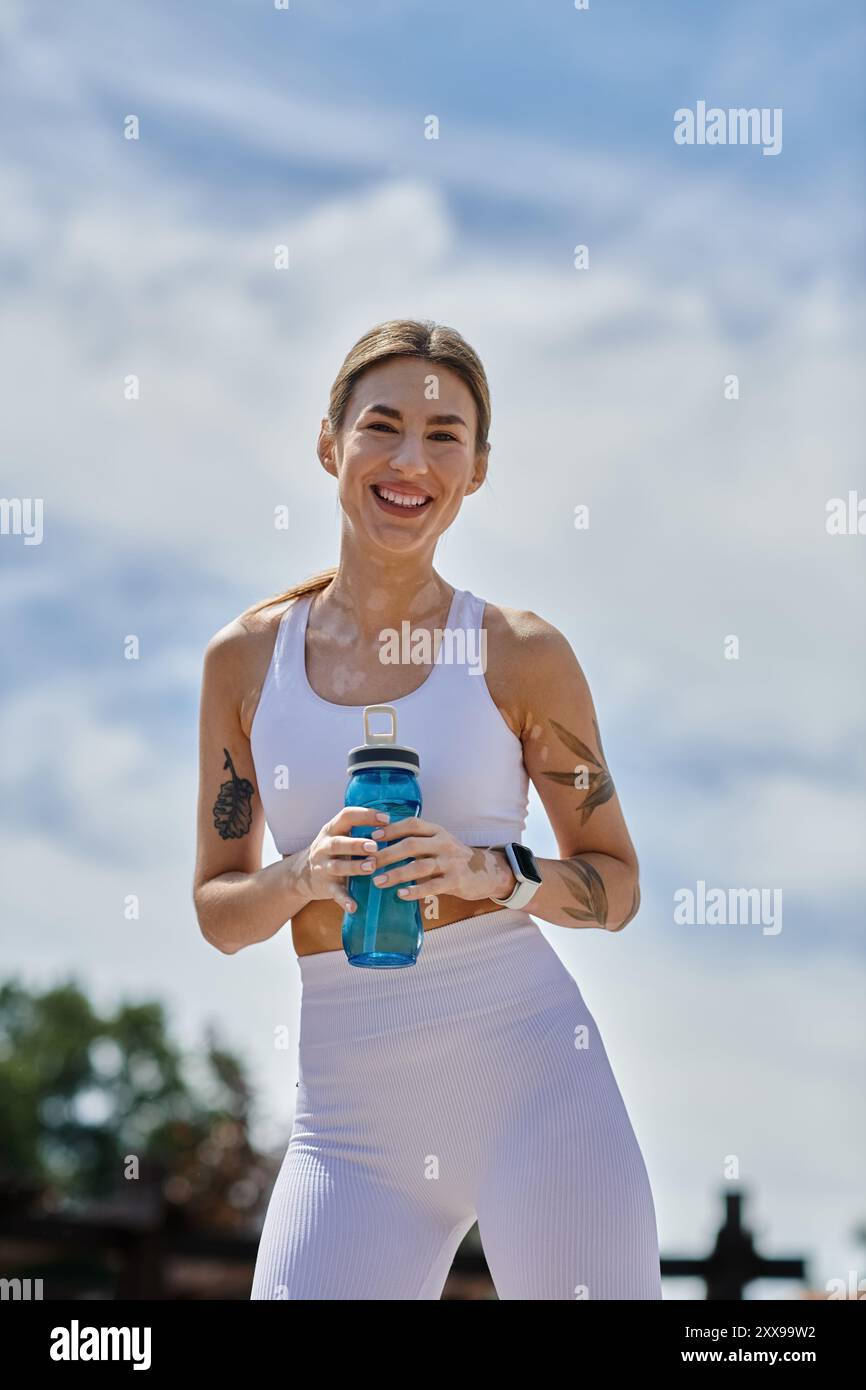 A young woman in white activewear smiles and holds a water bottle while ...