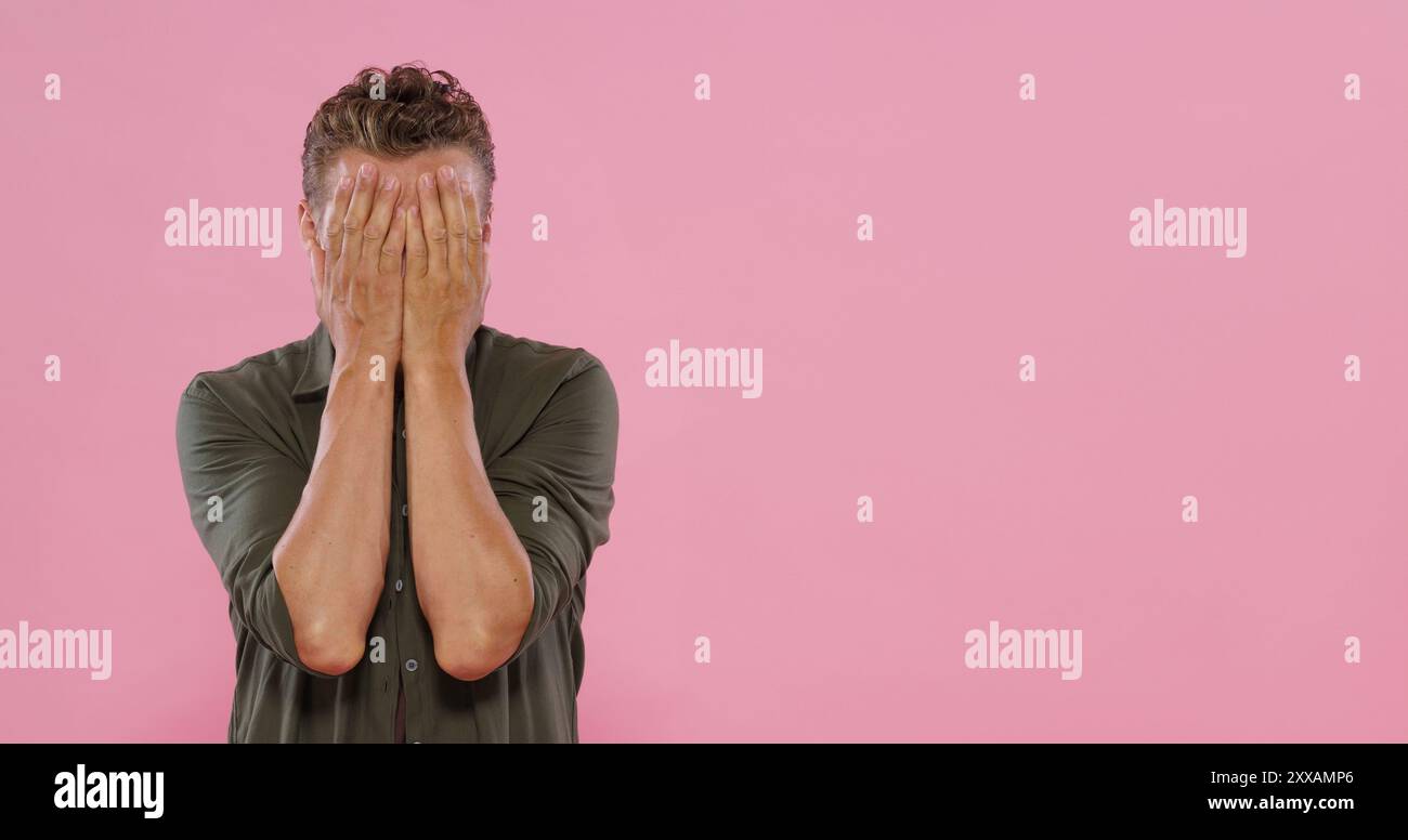 A man is standing in front of a pink wall, with his hands covering his ...