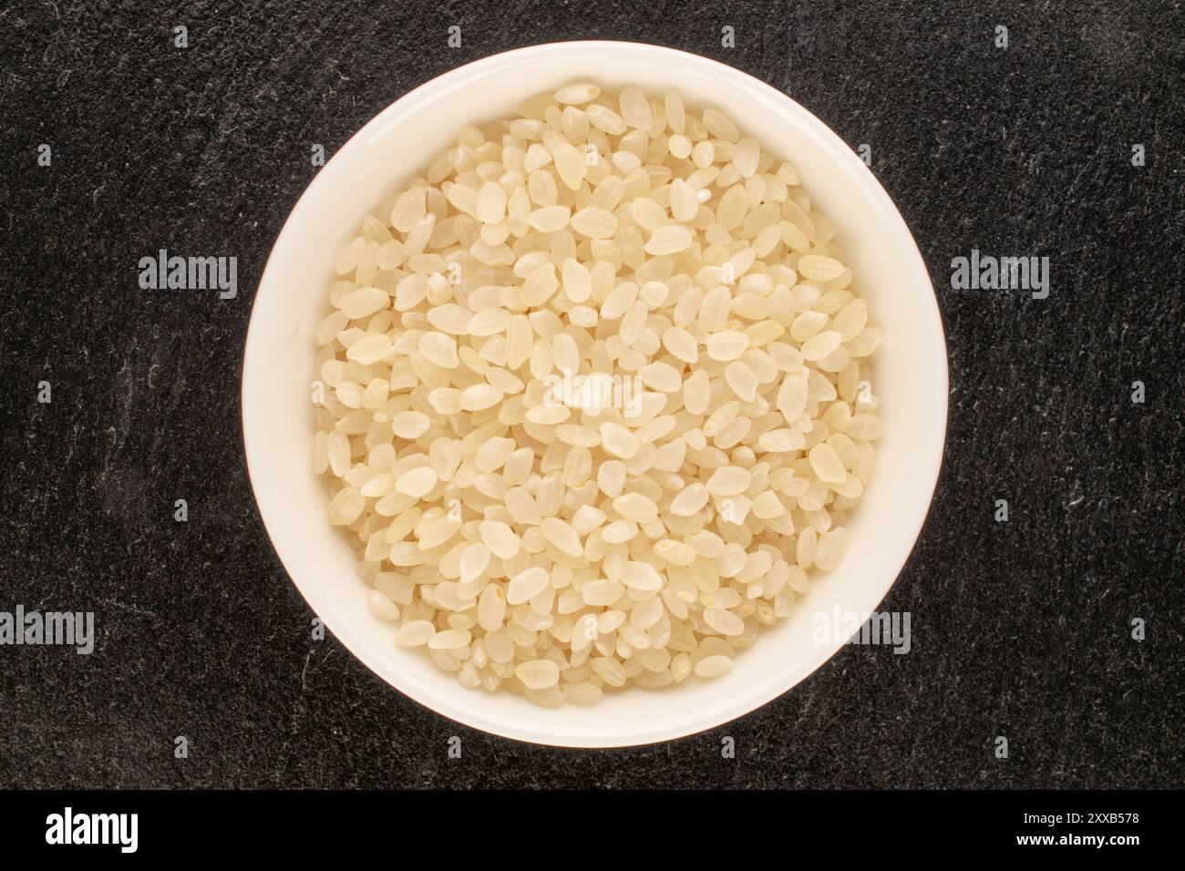 Round grain uncooked rice with white ceramic saucer on slate stone ...