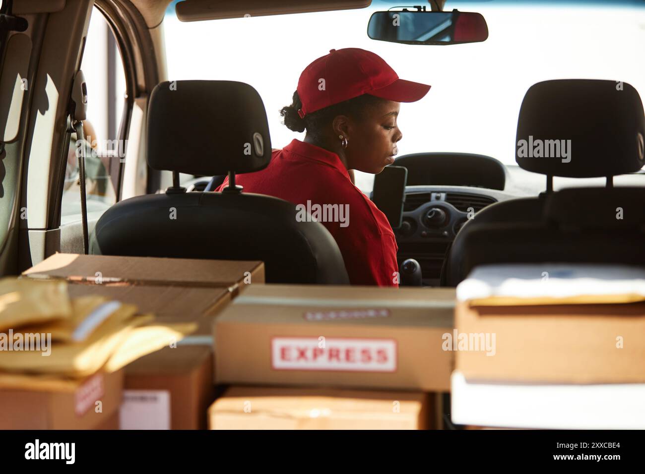 Worker in delivery uniform preparing packages inside vehicle, organizing boxes in back seat surrounded by parcels while focusing on task with express label in foreground Stock Photo
