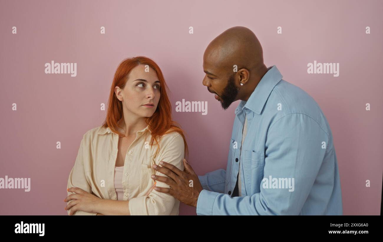 Interracial couple stands against a pink background with the man expressing emotions towards the woman who looks concerned and has her arms crossed em Stock Photo
