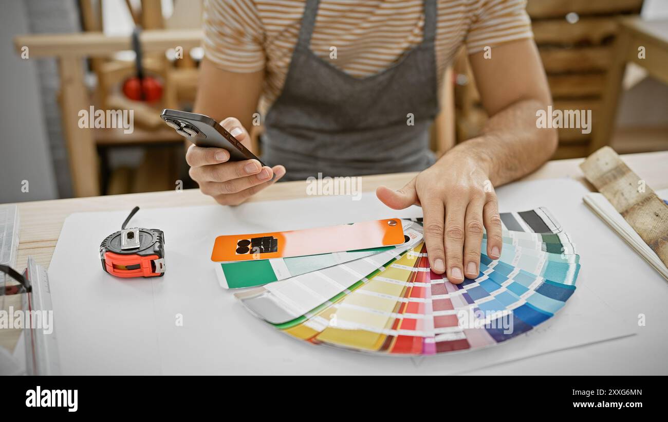 A young man examines color swatches in a workshop while using a smartphone, surrounded by carpentry tools. Stock Photo