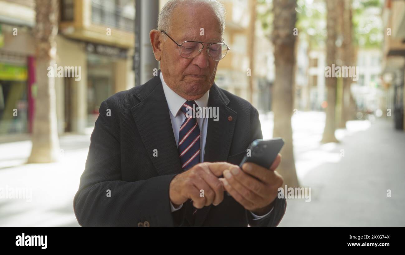 An elderly grey-haired caucasian man uses his smartphone while standing on an urban street during daylight. Stock Photo