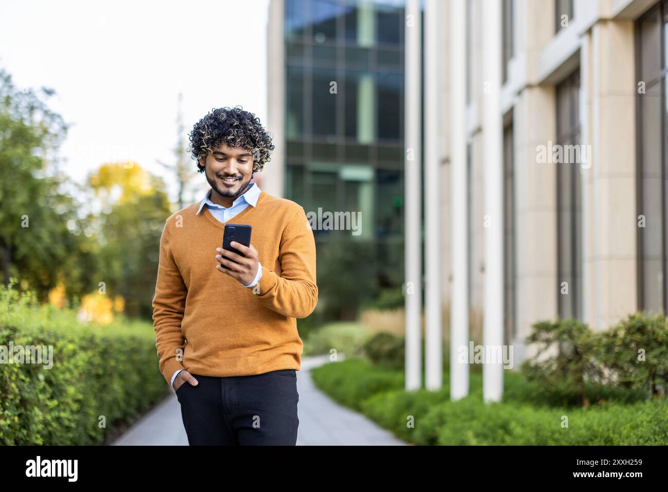 Young man in orange sweater uses phone while walking in urban area. Happy with technology interaction. Sunny day highlights blend of nature and architecture in modern city setting. Stock Photo