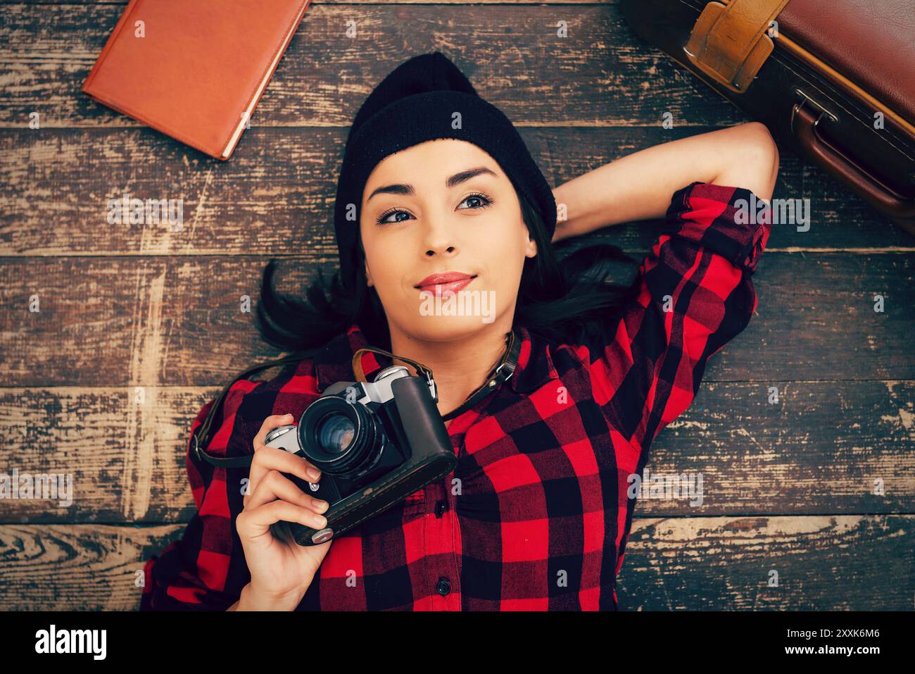 Beautiful day dreamer. Top view of beautiful young woman in headwear lying on the floor and holding camera while suitcase and note pad laying near her Stock Photo