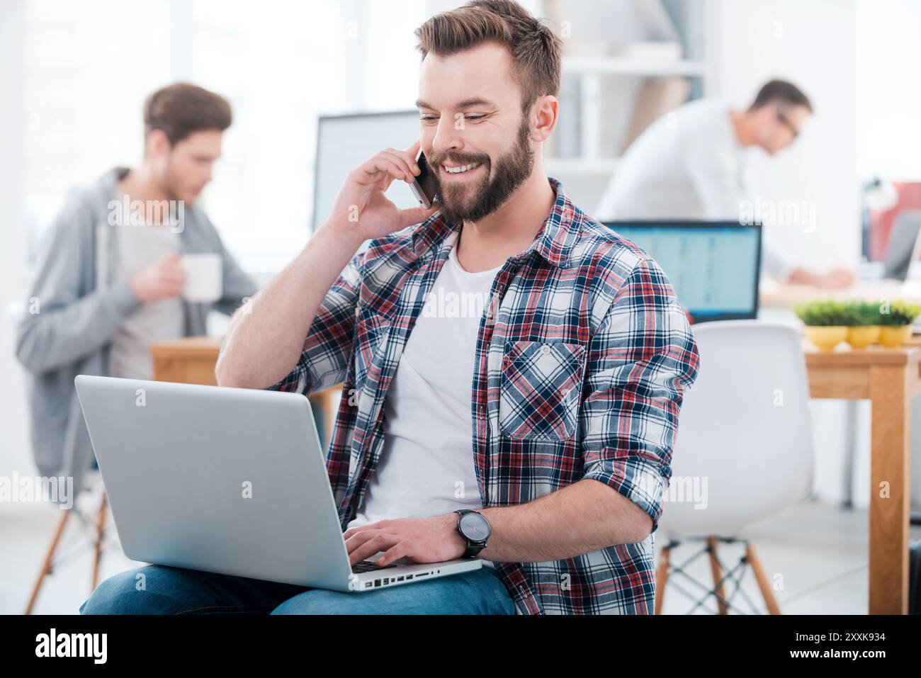 When the work is in full swing. Happy young man working on laptop and talking on the mobile phone while two people working in the background Stock Photo