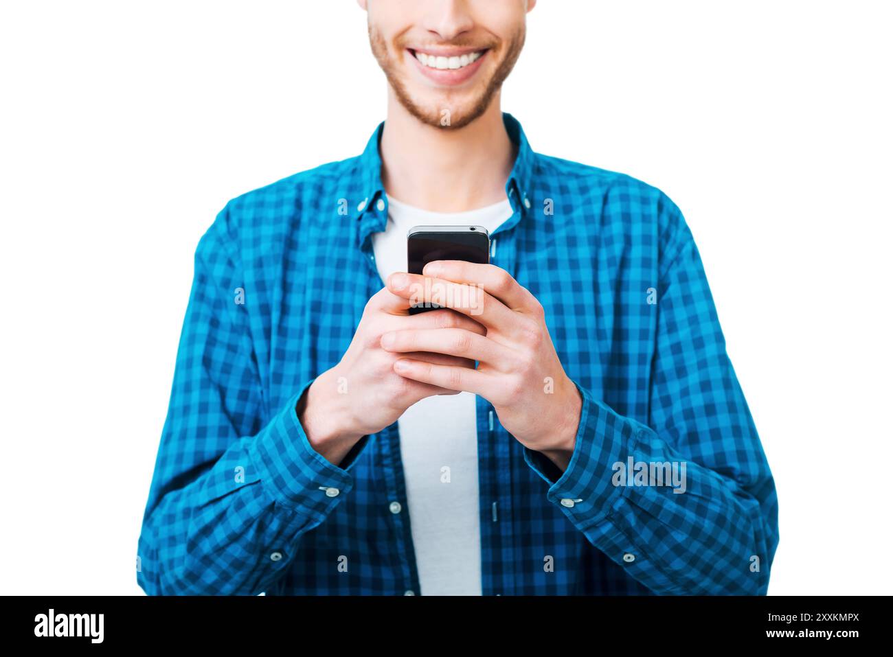 Choosing right words. Close-up of young man in shirt holding mobile phone and smiling while standing against white background Stock Photo