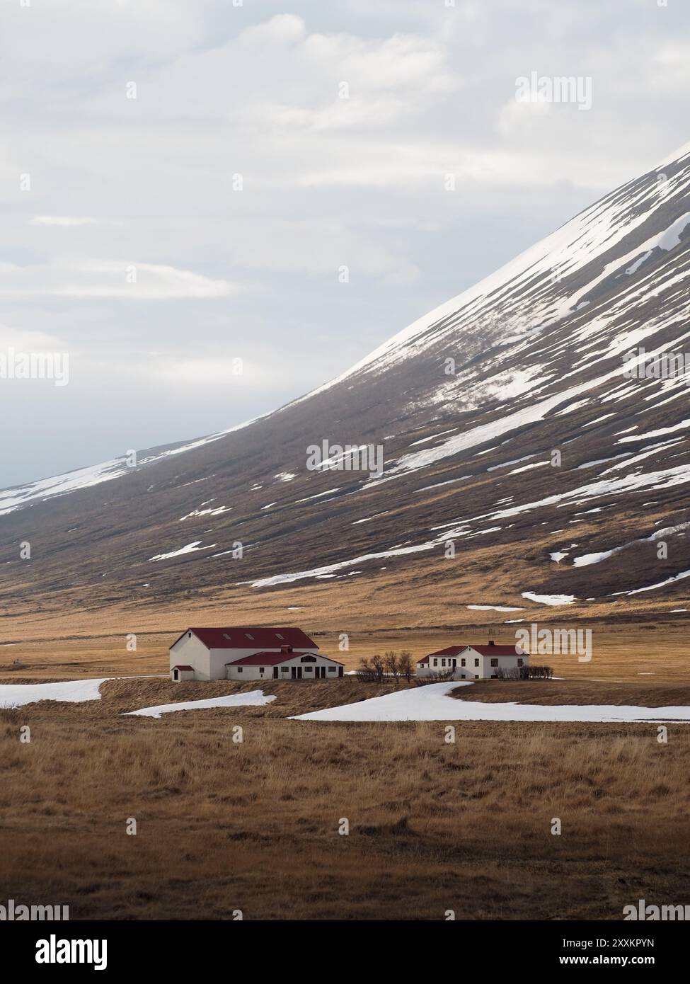 The photo depicts two quaint farmhouses near the snow-tinged foothills of a large mountain, with a cloud-strewn sky above, representing the quietude o Stock Photo