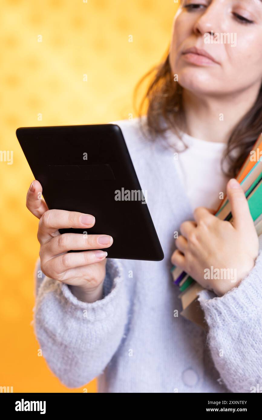 Woman carrying stack of books, reading online courses files for school exam on tablet, studio background. Student holding textbooks, doing research for university assessment on internet using device Stock Photo