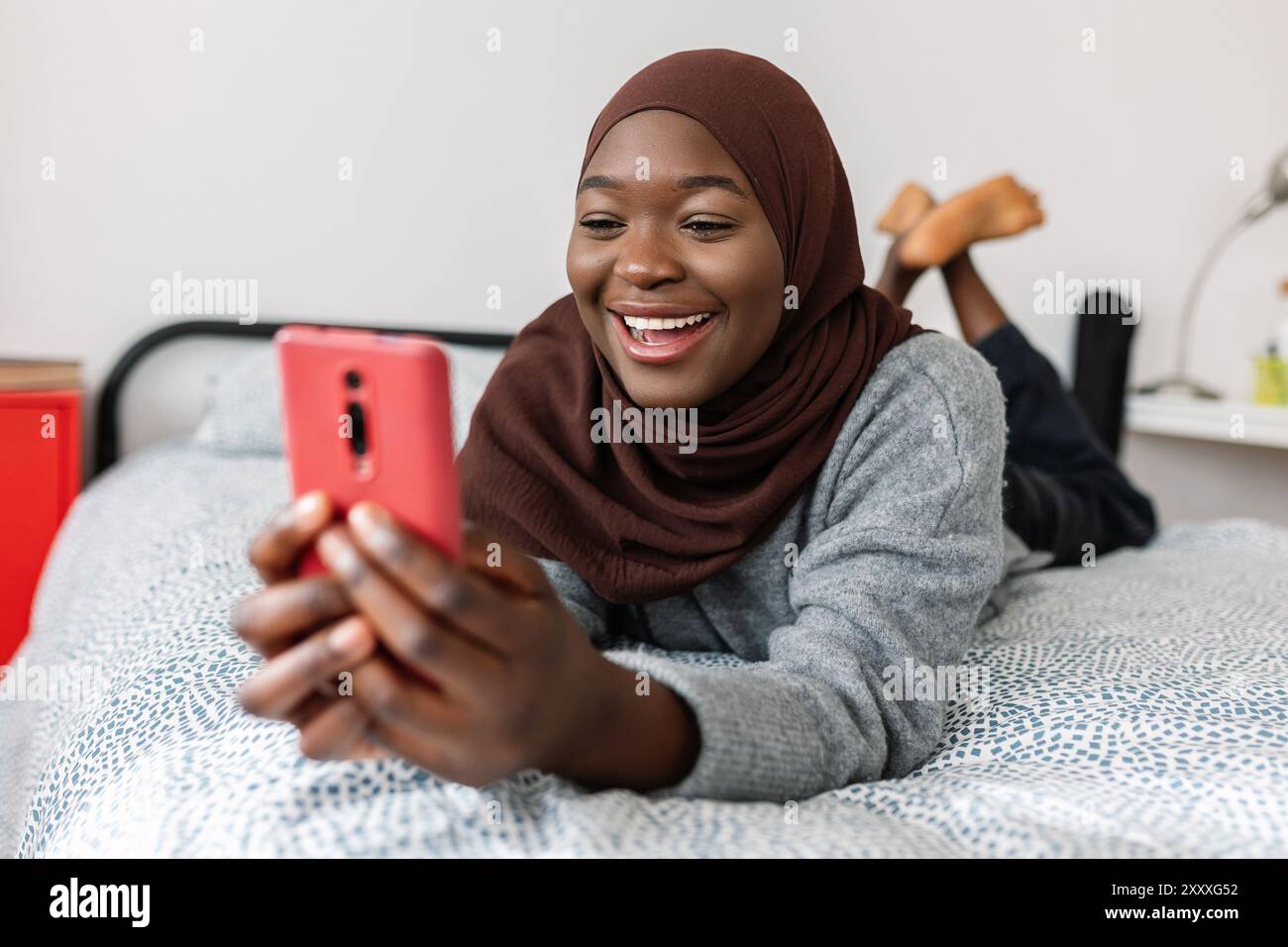 Happy black muslim woman in hijab using smartphone relaxing on bed at ...