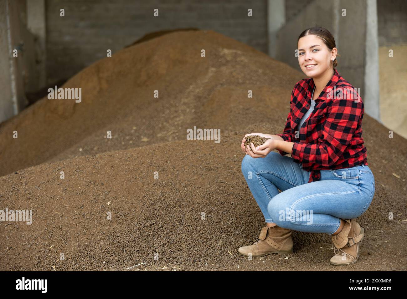 Fodder Worker Young dairy farm female worker squatting at heap of soybean husk in ...