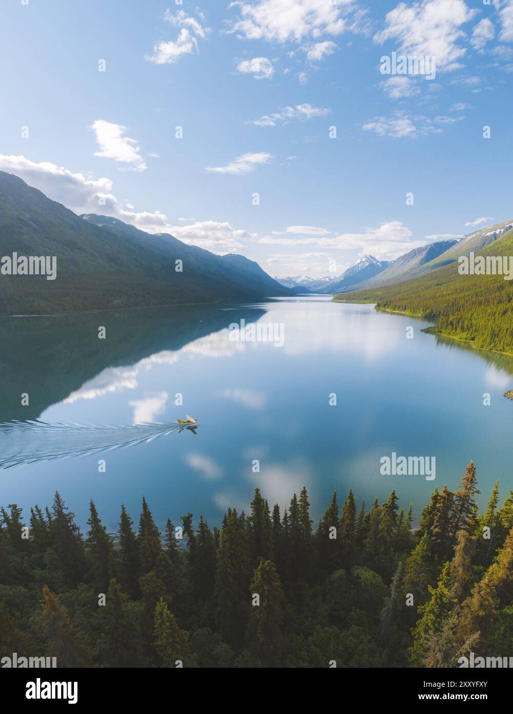 A tranquil lake reflecting the surrounding mountains and forest, with a small boat leaving a wake under a clear sky, Kusawa Lake, Yukon, Canada, North Stock Photo