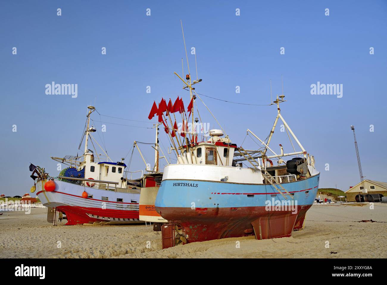 Fishing boats lying on the beach under a blue sky, some red flags waving in the wind, buildings in the background, Lokken, Jutland, Denmark, Europe Stock Photo