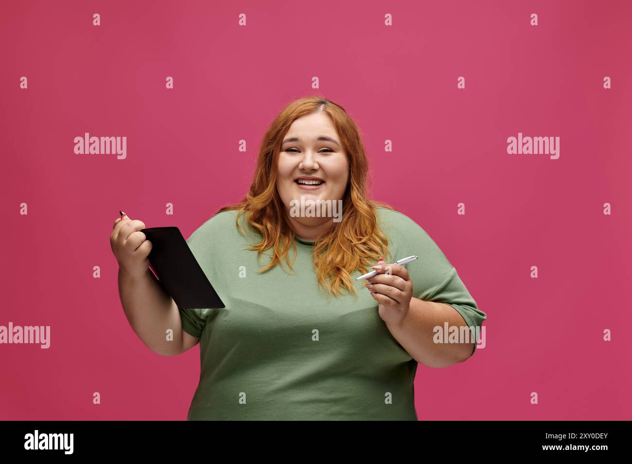 A plus size woman smiles while holding a notebook and pen in front of a pink background. Stock Photo